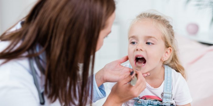 Sick child opening mouth while looking at doctor examining her sore throat with medical tool